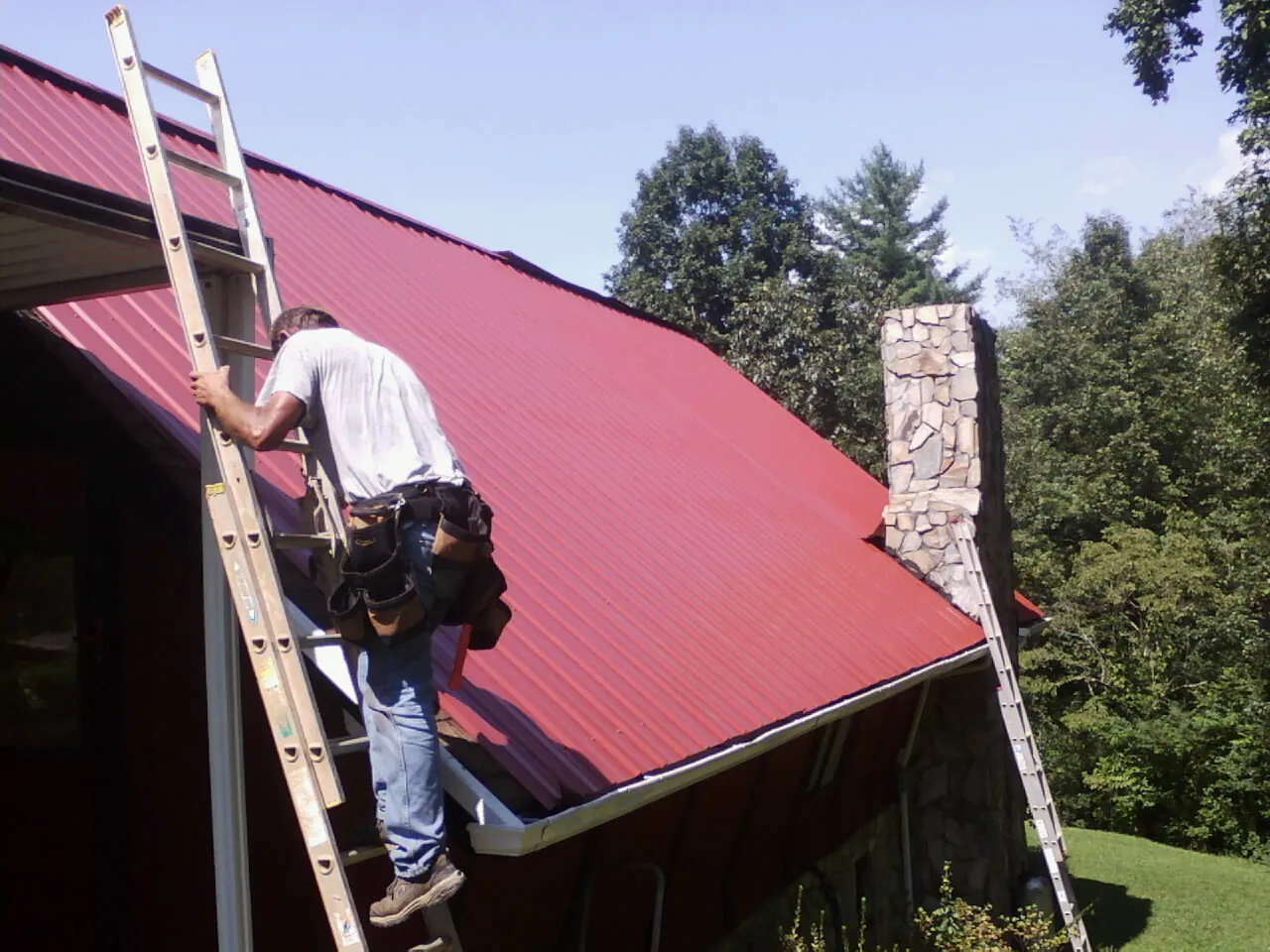 Roofer on a ladder beside a red metal roof and stone chimney — Soffit & Fascia Repair by Roofing Brothers in Alta Vista